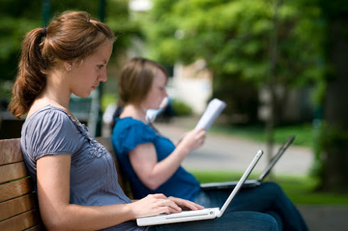two-girls-sitting-on-bench-with-laptops-open-while-one-is-reading-a-textbook-while-the-other-types-into-computer-outside